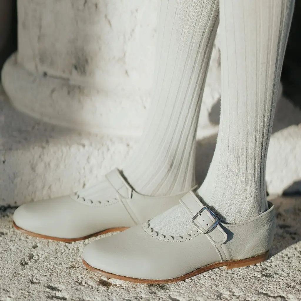 Close-up of feet wearing ribbed white tights and the Mini Mary Janes in Scalloped Pearl by Zimmerman Shoes, featuring a buckle, standing on a textured white surface.