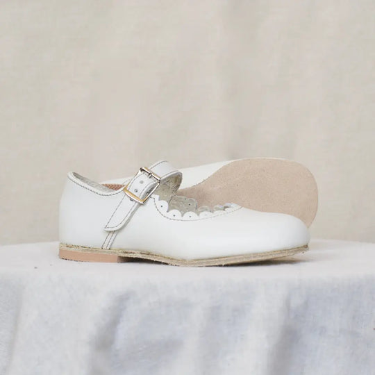 A pair of Mini Mary Janes in Scalloped Pearl by Zimmerman Shoes, featuring an adjustable ankle strap with buckle, is displayed on a light fabric surface against a neutral background.