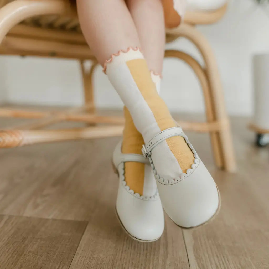 A child wearing Zimmerman Shoes’ Mini Mary Janes in Scalloped Pearl, featuring white leather and an adjustable ankle strap, sits on a wooden chair with their legs crossed, resting on a light wooden floor and wearing tall yellow-and-white socks.