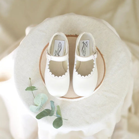 A pair of Zimmerman Shoes Mini Mary Janes in Scalloped Pearl, crafted from white leather with scalloped edges and an adjustable ankle strap, are displayed on a round wooden surface with a small sprig of green leaves beside them.