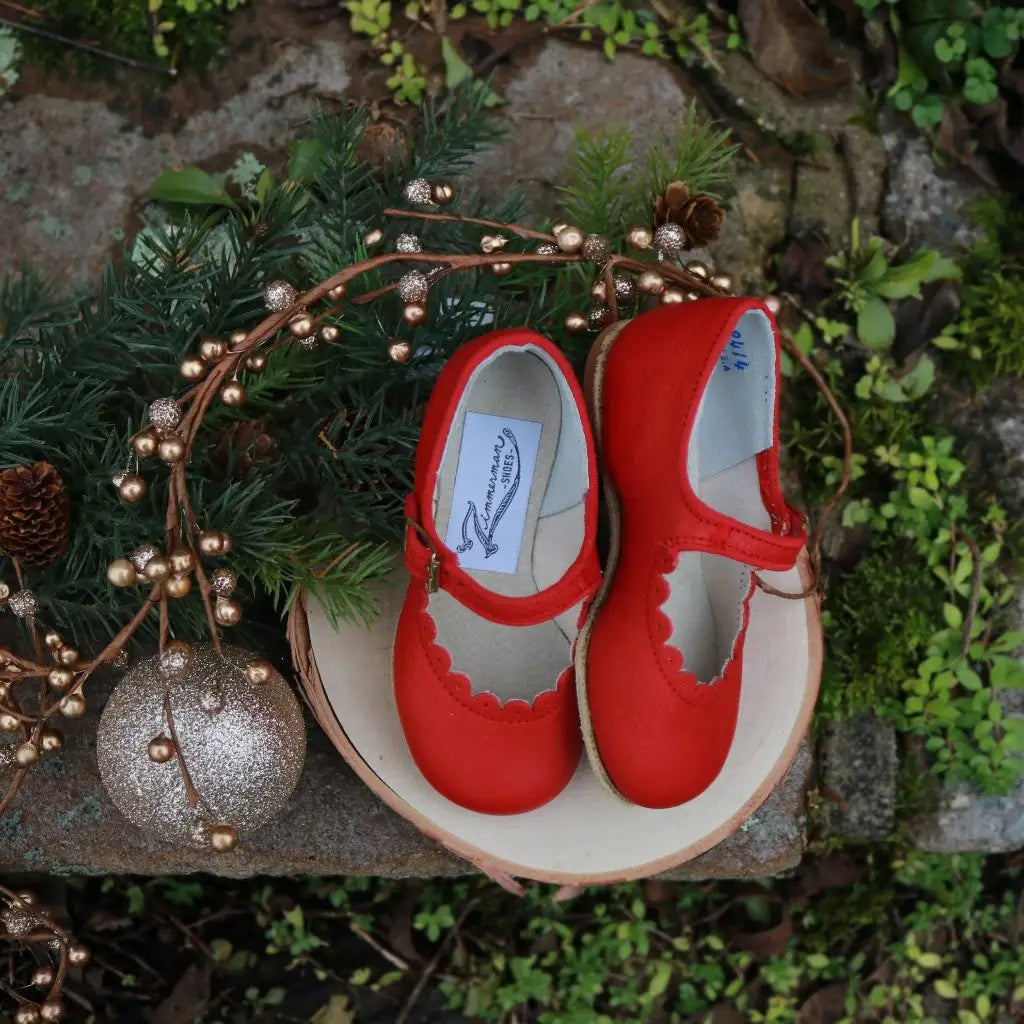 The Mini Mary Janes in Scalloped Red by Zimmerman Shoes, a pair of small red leather children’s shoes with scalloped edges and adjustable ankle straps, rest on a wooden slice surrounded by pine branches, gold beads, and a glittery gold ornament outdoors.