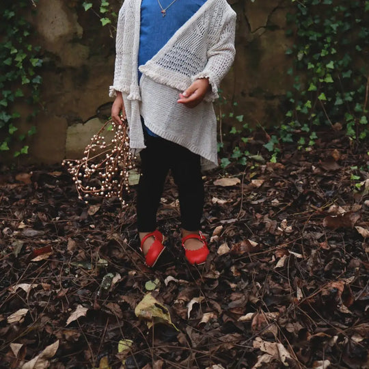 A child in a blue dress, white cardigan, and black leggings stands on dry leaves, holding string lights. They are wearing Zimmerman Shoes’ Mini Mary Janes in Scalloped Red—red leather shoes featuring an adjustable ankle strap and scalloped edge. Lush green ivy climbs the stone wall behind them, and the child’s face is not visible.