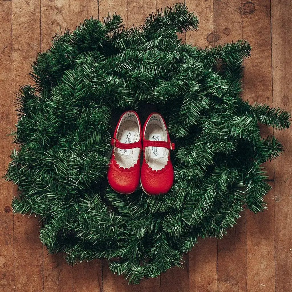 A pair of Zimmerman Shoes Mini Mary Janes in Scalloped Red, featuring a charming scalloped edge and adjustable ankle strap, are displayed atop a green artificial Christmas wreath on a wooden floor.