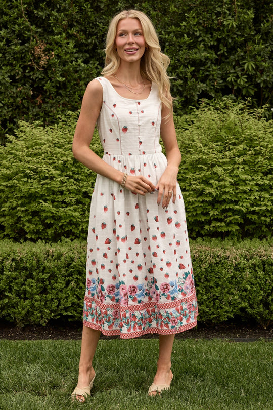 A smiling woman stands on grass, lush green bushes behind her, wearing the Ivy City Co Sadie Dress in Strawberry Vintage Tier—a sleeveless white cotton summer dress adorned with red and pink floral patterns. She has long blonde hair and beige shoes.