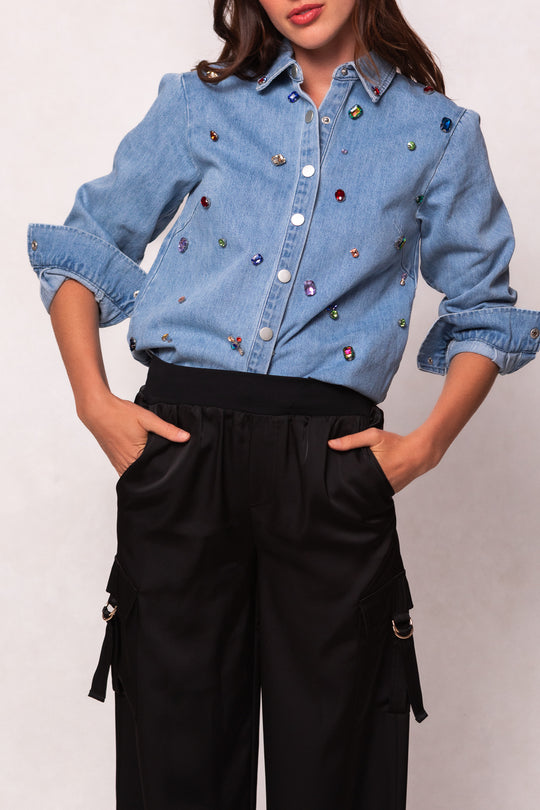 Person wearing a blue denim shirt with colorful embellishments and black pants on a white background