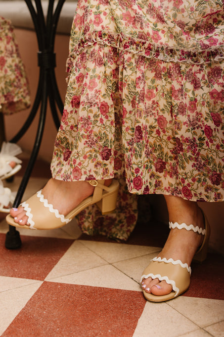 Seated indoors on a red and white tiled floor, a person wears Ivy City Co’s Lois Heel in Tan, featuring adjustable ankle straps, paired with a long floral dress.