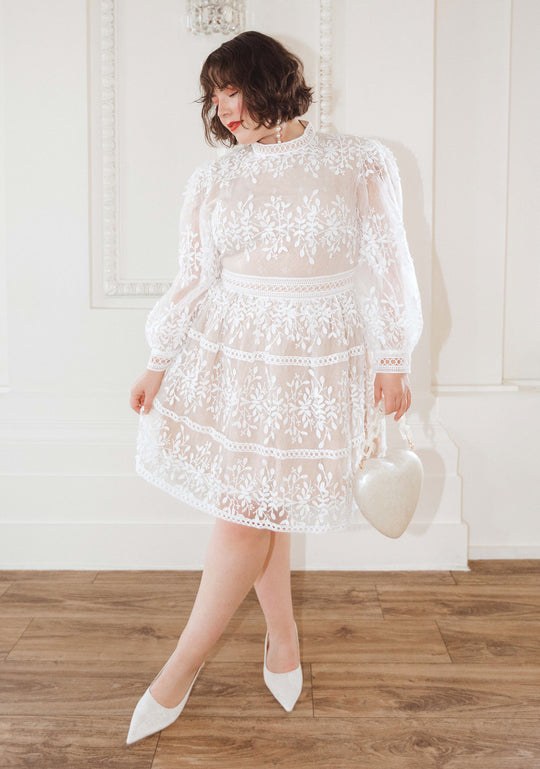 A woman in an Ivy City Co Sicily Short Dress poses indoors, holding a heart-shaped white handbag and wearing white heels, highlighting the elegant womens dress against crisp white walls and wooden flooring.