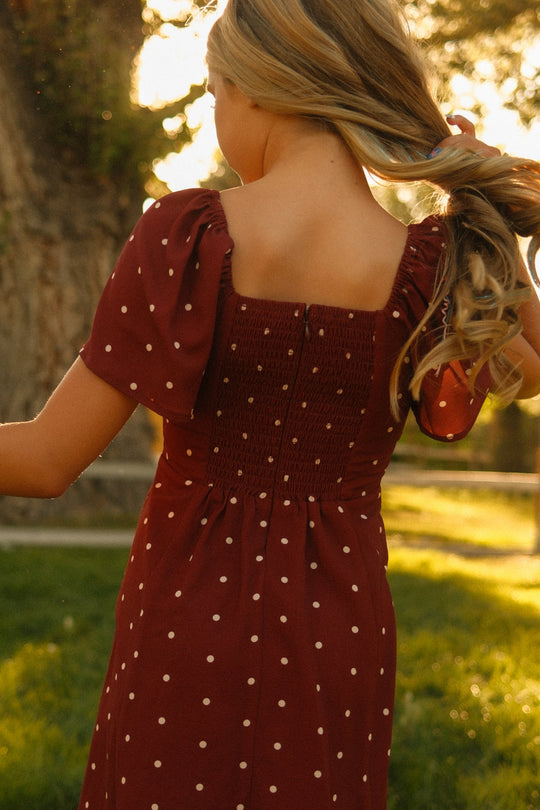 A person with long blonde hair, wearing the Ivy City Co Teen Blakely Dress in Cranberry Dainty Dot—a red, midi-length dress with white polka dots and petal sleeves—stands outside holding their hair as sunlight filters through the trees in the background.