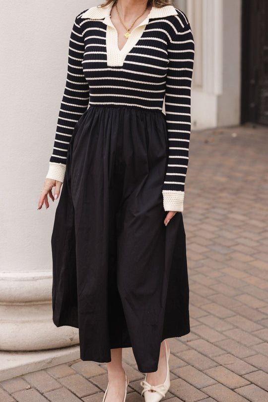 A woman stands on a brick sidewalk wearing the Adelina Dress in Black by Ivy City Co, featuring a striped knit bodice and cream collar. She completes the look with cream heels and a gold necklace. Her face is not visible.