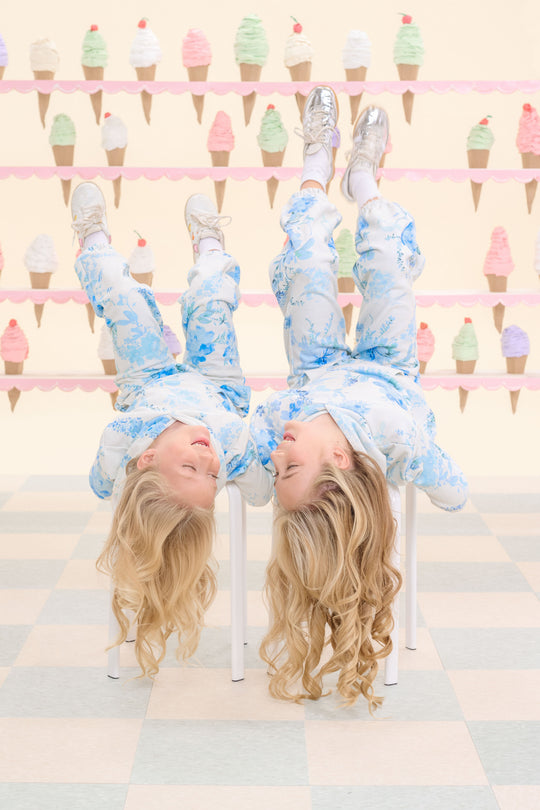 Two young girls with long blonde hair, dressed in matching blue and white pajamas and the Ivy City Co Mini Kian Hooded Sweatshirt in Blue Aquarelle Fleur, lie upside down on chairs. Behind them, pastel walls decorated with ice cream cone motifs create a whimsical backdrop, while the floor features a light checkered pattern.