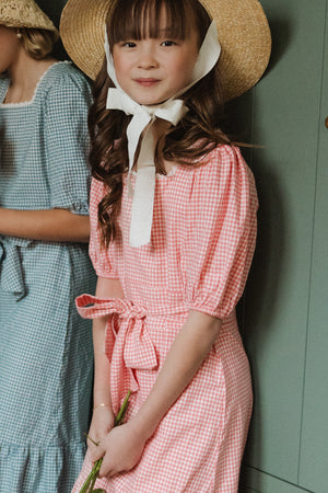 A young girl wears the Mini Hannah Dress in Pink Gingham by Ivy City Co, featuring a bow at the waist and a wide-brimmed straw hat tied under her chin, smiling softly. Another child in blue gingham stands beside her.