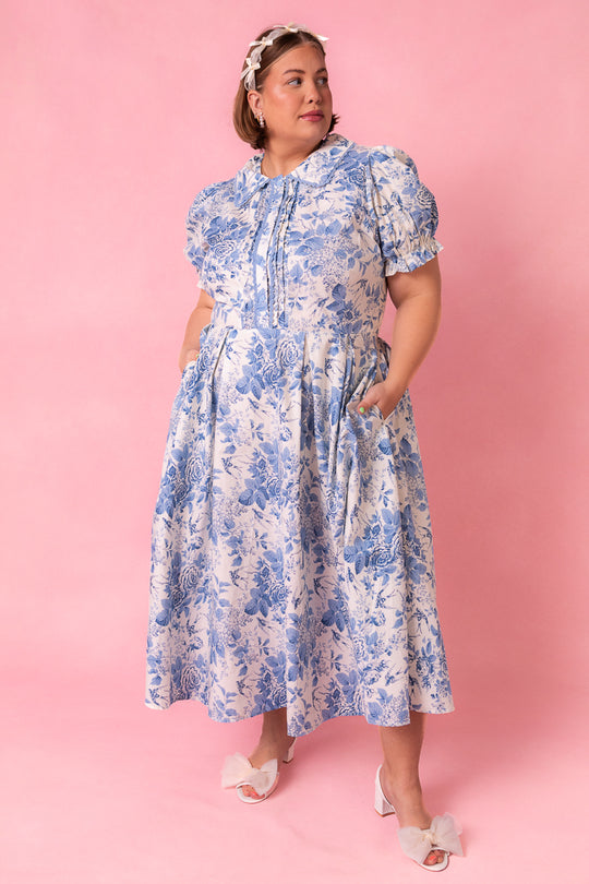A woman models the Ivy City Co Sophia Dress in Blue Floral—featuring puff sleeves, ruffle details, and nursing-friendly design—against a pink background. She accessorizes with a headband and white heeled sandals with bows, hands in her pockets.
