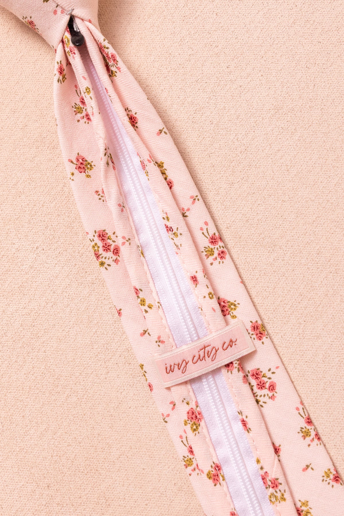 A close-up of the Ivy City Co Ties in Madeline Pink Floral, featuring a white center zipper and branded fabric label, laid flat on a soft peach background—ideal for matching family accessories.