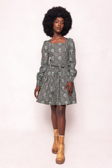 A woman with a natural afro hairstyle wears the Ivy City Co Finn Dress in Garden Tile—a vintage-inspired green floral long-sleeve dress with a square neckline—and brown ankle boots, standing against a light background.