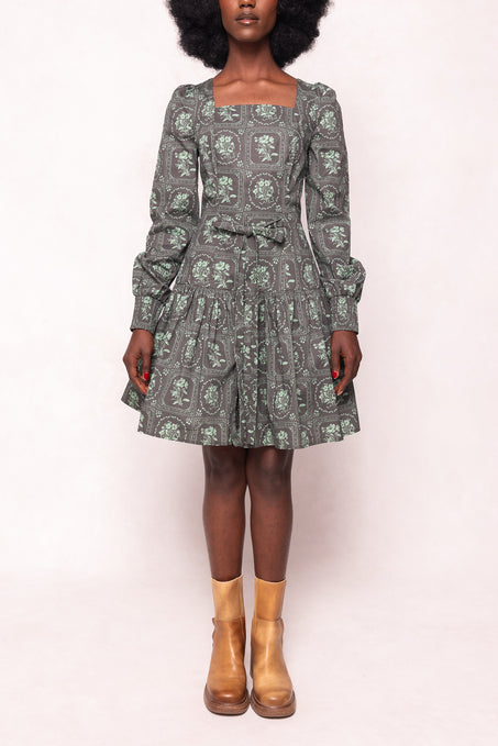 A woman wears the Finn Dress in Garden Tile by Ivy City Co, featuring a vintage-inspired green floral print, long sleeves, a square neckline, and a tiered skirt. She pairs it with tan ankle boots and stands against a light background, her face partially out of frame.