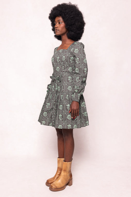 A woman with natural afro hair stands sideways wearing the Ivy City Co Finn Dress in Garden Tile, a vintage-inspired green floral long-sleeve dress with a flared skirt, paired with brown ankle boots against a plain light background.