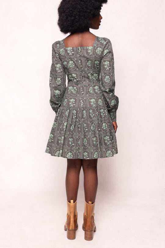 A woman with an afro faces away, wearing Ivy City Cos Finn Dress in Garden Tile—a vintage-inspired green floral long sleeve dress with a square neckline and pleated skirt—paired with tan heeled boots against a light background.