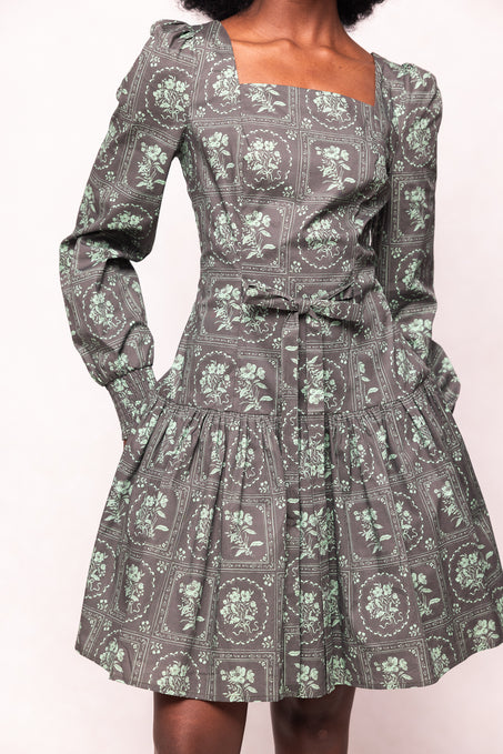 A woman wears the Ivy City Co Finn Dress in Garden Tile—a vintage-inspired green floral print, long-sleeve, square-neck gray dress with a fitted waist, pleated skirt, and pockets. Her hands are in the pockets and her head is not visible.