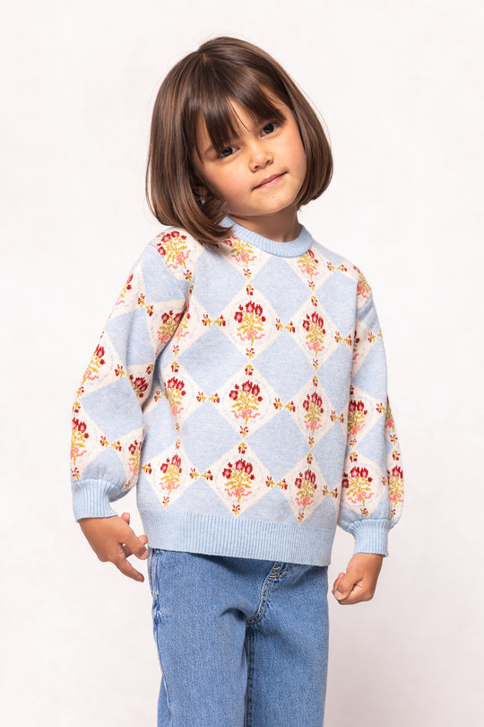 A young child with straight brown hair wears the Ivy City Co Mini Vivienne Sweater in Mosaic Bloom, featuring diamond and floral patterns in baby blue, paired with light blue jeans against a plain white background.