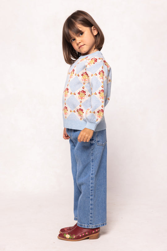 A young girl stands sideways against a plain light background, wearing the Ivy City Co Mini Vivienne Sweater in Mosaic Bloom, paired with loose blue jeans and red floral-detailed shoes.