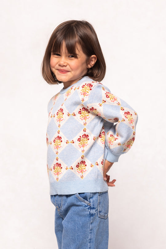 A young girl with short brown hair smiles playfully in the Ivy City Co Mini Vivienne Sweater in Mosaic Bloom and blue jeans, standing against a plain light background.