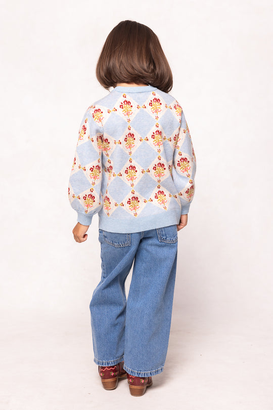 A young child with shoulder-length brown hair faces away, wearing the Ivy City Co Mini Vivienne Sweater in Mosaic Bloom—a baby blue girls’ sweater with a floral diamond pattern—paired with blue wide-leg jeans and brown shoes against a plain background.