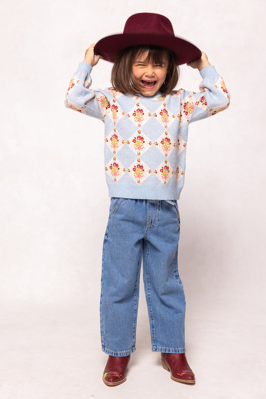 A young child smiles while playfully holding a maroon hat, wearing the Ivy City Co Mini Vivienne Sweater in Mosaic Bloom, loose blue jeans, and matching maroon boots, standing against a light background.