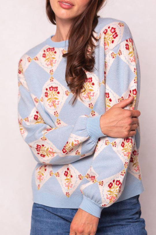 A woman with long brown hair wears the Ivy City Co Vivienne Sweater in Mosaic Bloom, a light blue maternity-friendly piece with colorful geometric and floral patterns, paired with blue jeans. She poses with arms crossed and her face partially visible.