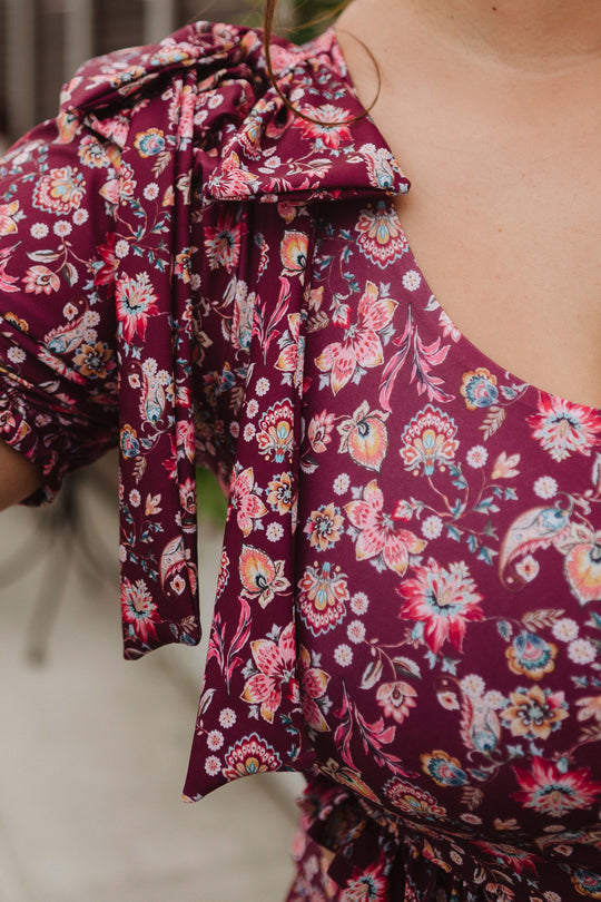 A close-up of a person wearing the Ivy City Co Sailor Top in Stephanie Floral, featuring a maroon base with colorful florals, a bow on the shoulder, gathered puff sleeves, and a rounded neckline against a blurred background.