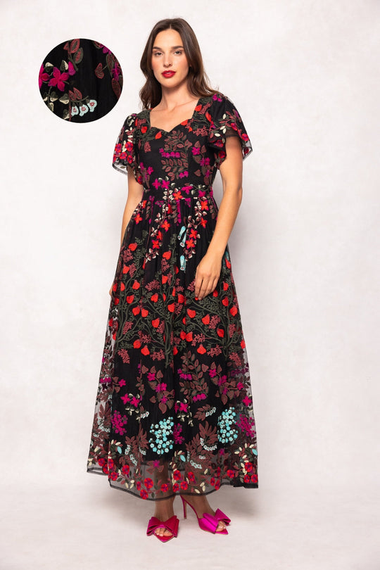 A woman wears the Flora Dress in Moonlit Blossom by Ivy City Co—a black, floor-length gown adorned with vibrant, intricately embroidered florals. The dress features short flutter sleeves and a belted waist. She stands against a light background, and there is a close-up detail showcasing the elaborate embroidery on the fabric.