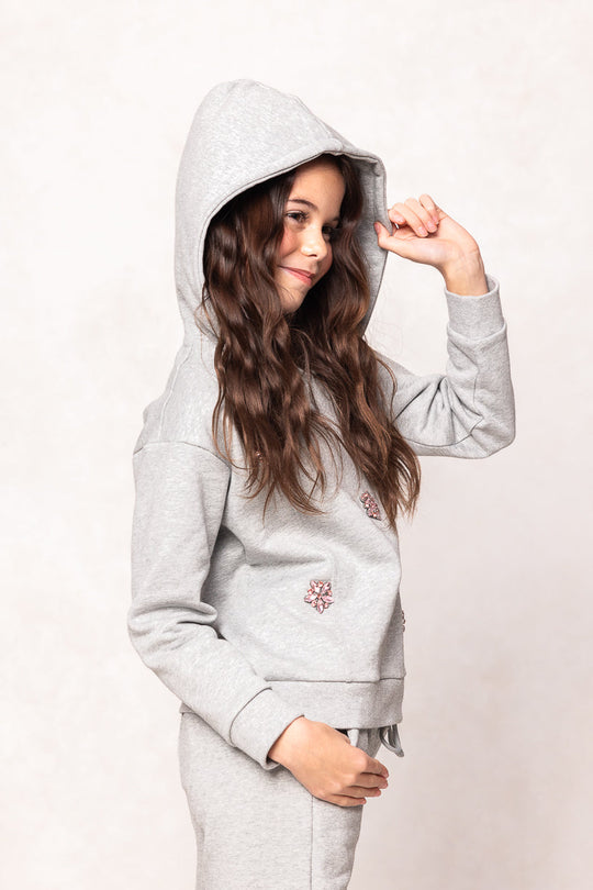 A young girl with long brown hair smiles and holds up her hood while wearing the Ivy City Co Mini Skylar Hoodie in Heather Gray, set against a light background.