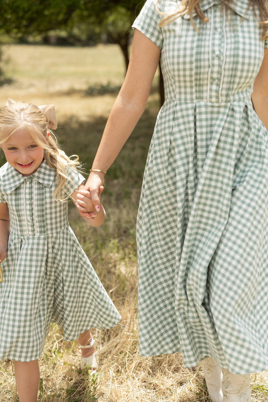 A woman and a young girl, both smiling, hold hands while walking outdoors on dry grass, wearing matching Ivy City Co Mini Amber Dresses in Sage Gingham with pleated skirts.