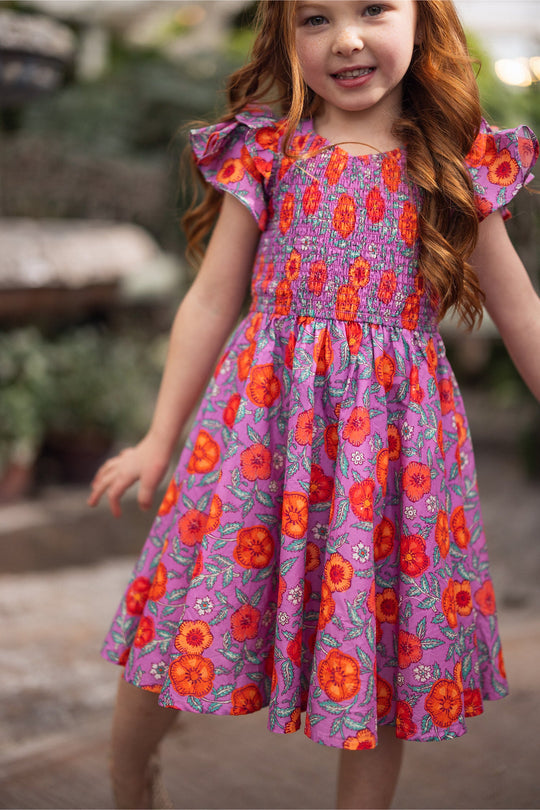 A young girl with long red hair wears the Mini Hattie Dress in Thistle Flower Poppy Woodblock by Ivy City Co, featuring a vibrant purple floral pattern with bright orange and red flowers, as she smiles outdoors surrounded by greenery.