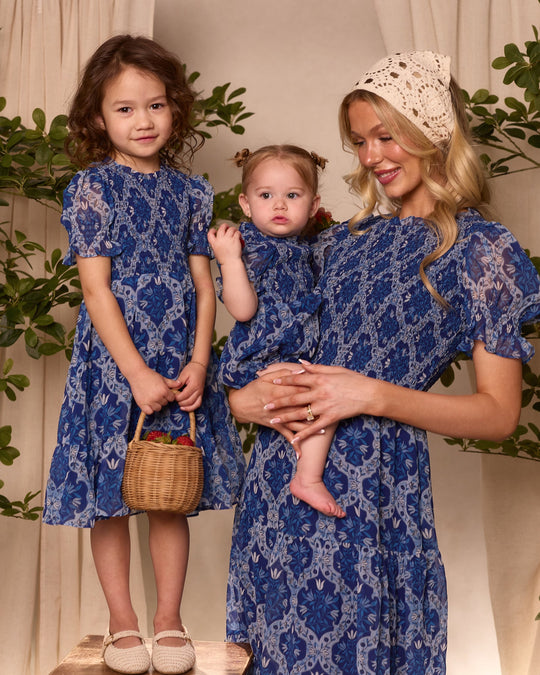 A smiling woman holding a toddler stands beside a young girl carrying a basket. All are wearing matching family outfits from Ivy City Co, including the Mini Cordelia Dress in Trellis Foulard and girls’ chiffon dress in beautiful blue patterns. The backdrop is decorated with lush green plants and beige curtains.