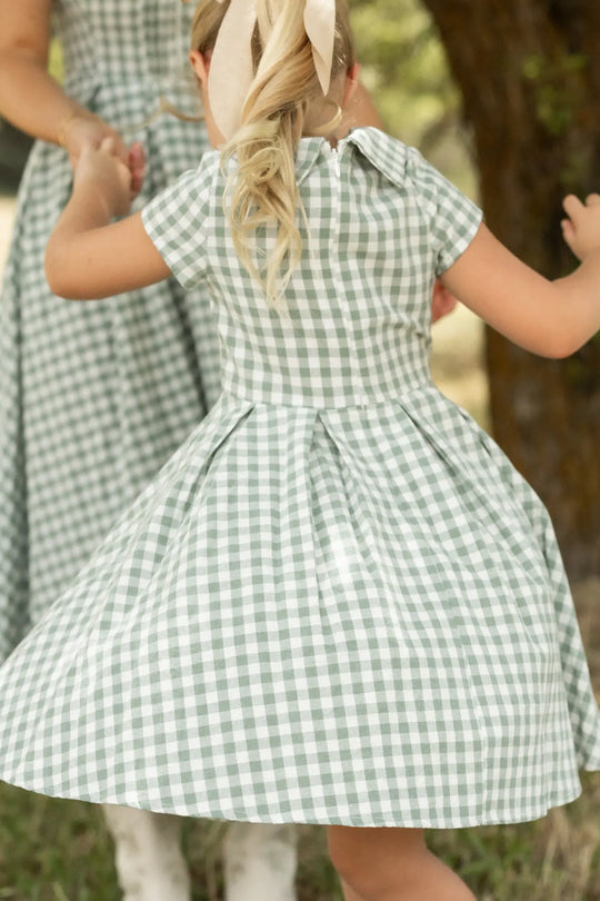 A young girl wearing the Ivy City Co Mini Amber Dress in Sage Gingham, featuring a pleated skirt, twirls outside while holding hands with an adult dressed similarly. Her blonde hair is tied back with a light-colored ribbon.