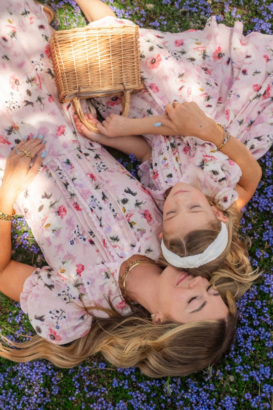 Two women in matching Ivy City Co Sophia Dress in Roselyn Pink lie on grass sprinkled with small purple flowers, heads together and eyes closed, looking relaxed and peaceful as they hold a wicker basket.