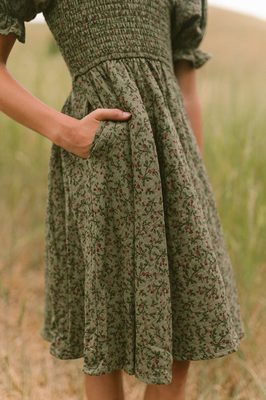 A person wearing the Ivy City Co Mini Teagan Dress in Woodland Floral, featuring short sleeves and a smocked bodice, stands outdoors with one hand in a pocket as the green floral pattern blends into the grassy background.