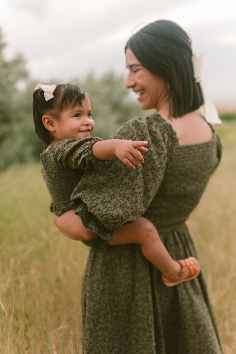 A woman in a green Woodland Floral dress smiles while holding a young girl wearing the Ivy City Co Baby Teagan Romper in Woodland Floral. Both have hair bows and look happy together in a grassy outdoor setting.