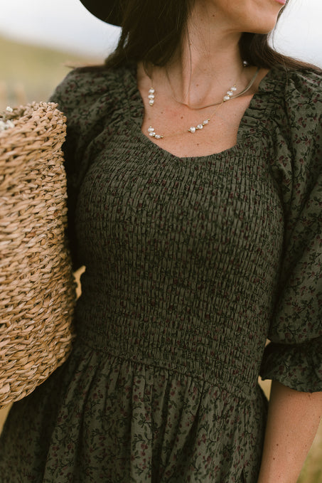 A woman with long brown hair, partially visible face, wears the Ivy City Co Teagan Dress in Woodland Floral and a pearl necklace while holding a woven straw basket.