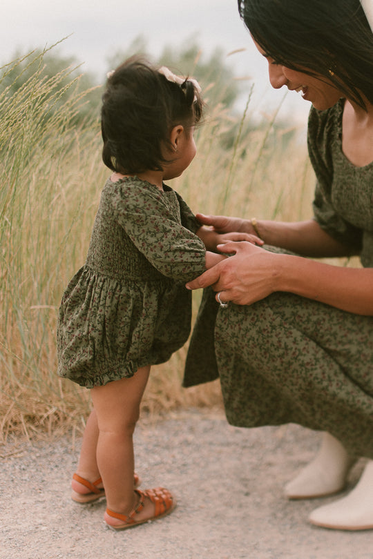 A toddler wearing the Ivy City Co Baby Teagan Romper in Woodland Floral stands holding hands with a smiling woman in matching attire, outdoors near tall grass.