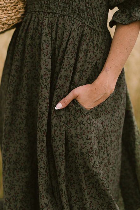 A person with light skin rests their hand in the pocket of the Ivy City Co Teagan Dress in Woodland Floral, which features a smocked bodice. The photo focuses on the lower torso and hand, with a woven basket partly visible in the background.