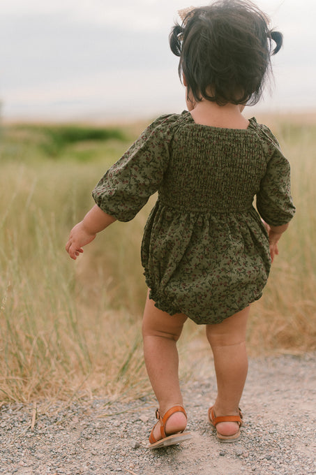 A toddler with dark hair walks away on a dirt path through grassy fields, wearing the Ivy City Co Baby Teagan Romper in Woodland Floral and brown sandals.