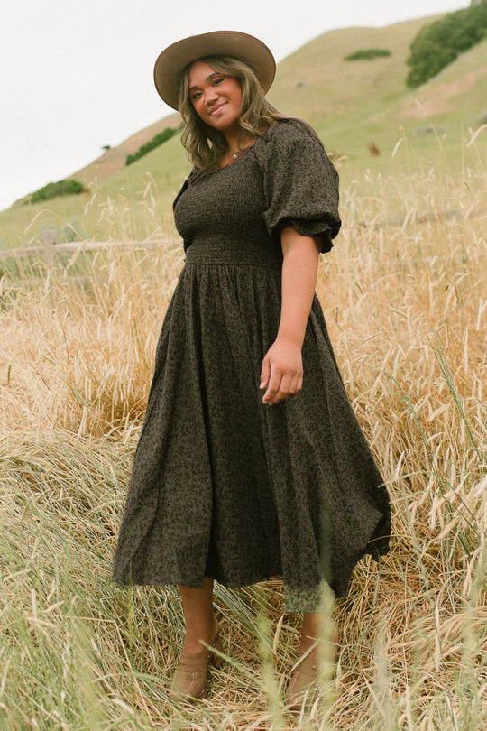 A woman in the Ivy City Co Teagan Dress in Woodland Floral, featuring a smocked bodice and wide-brim hat, stands smiling in a grassy field with hills behind her. The dress is maternity and nursing friendly.