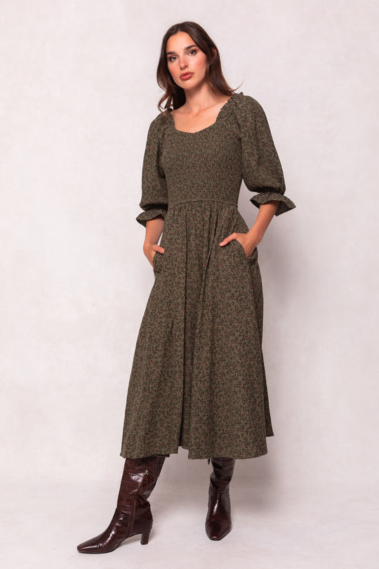 A woman wears Ivy City Co’s Teagan Dress in Woodland Floral—an olive green, long dress with puffed three-quarter sleeves, smocked bodice, and pockets—paired with brown knee-high boots against a light background.