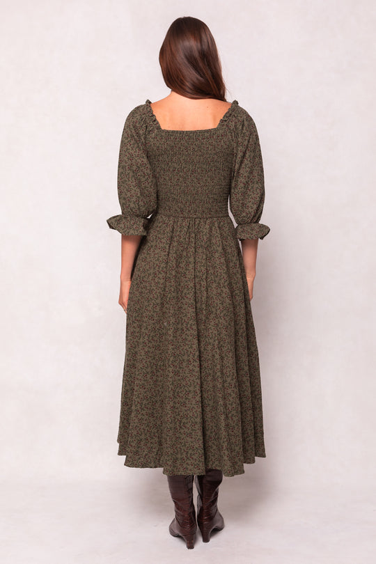 A woman with long brown hair, seen from behind, wears the Ivy City Co Teagan Dress in Woodland Floral—featuring puffy three-quarter sleeves and a smocked bodice, maternity nursing friendly—with brown boots against a light neutral background.
