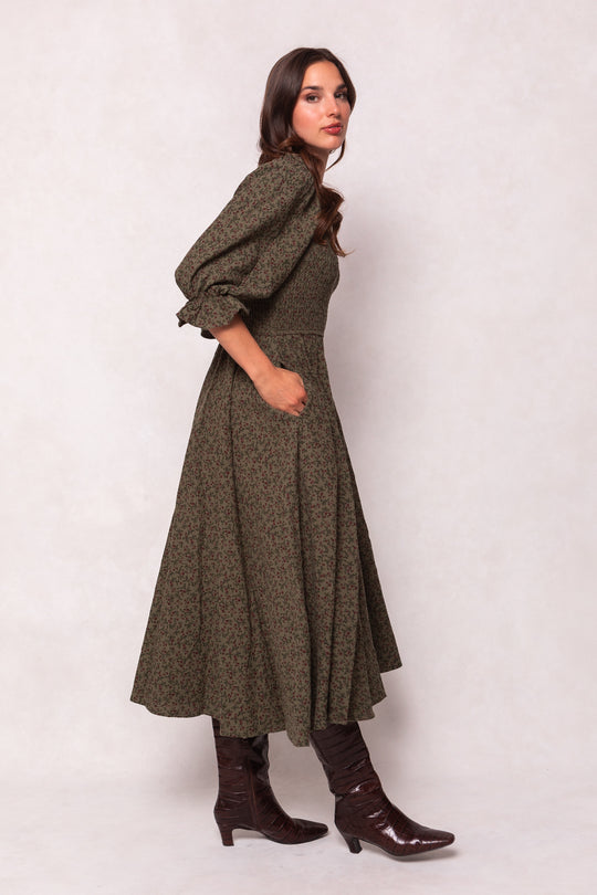A woman poses sideways against a neutral background in the Teagan Dress in Woodland Floral by Ivy City Co, featuring a long olive green design with a smocked bodice, puffed sleeves, and paired with brown heeled boots.