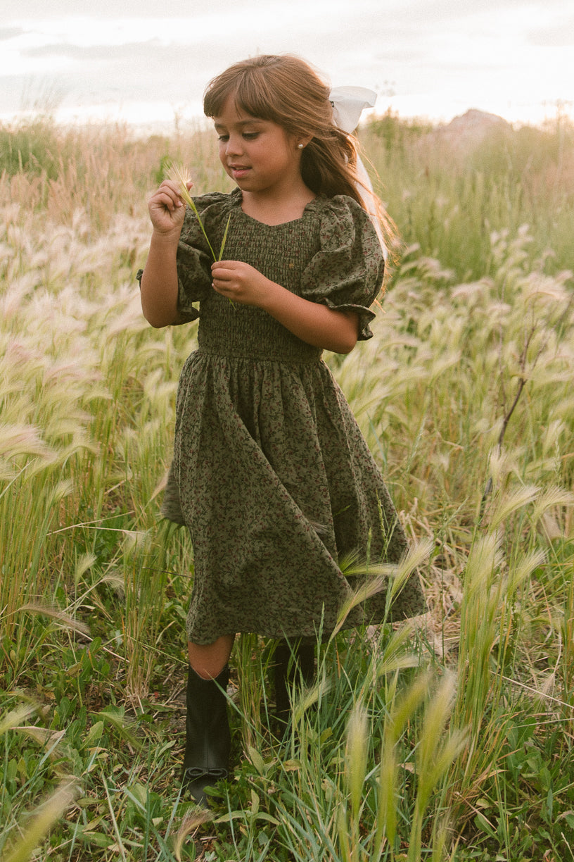 A young girl wears the Ivy City Co Mini Teagan Dress in Woodland Floral with black boots and a white bow, standing in a grassy field holding a flower under soft sunlight.