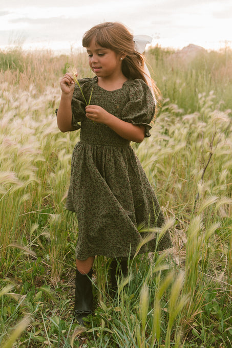 A young girl wears the Ivy City Co Mini Teagan Dress in Woodland Floral with black boots and a white bow, standing in a grassy field holding a flower under soft sunlight.