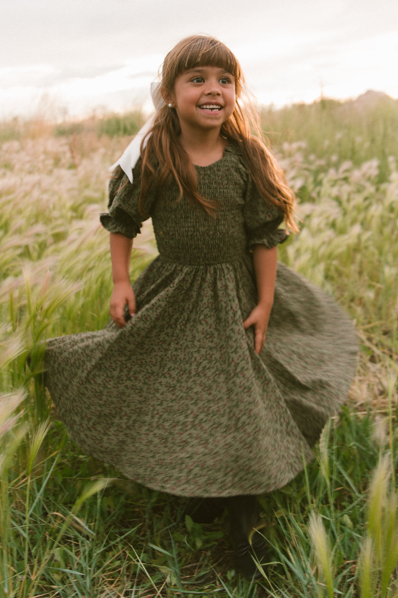 A young girl with long brown hair in a ribbon smiles and spins in the Ivy City Co Mini Teagan Dress in Woodland Floral among tall grass in a sunlit field.