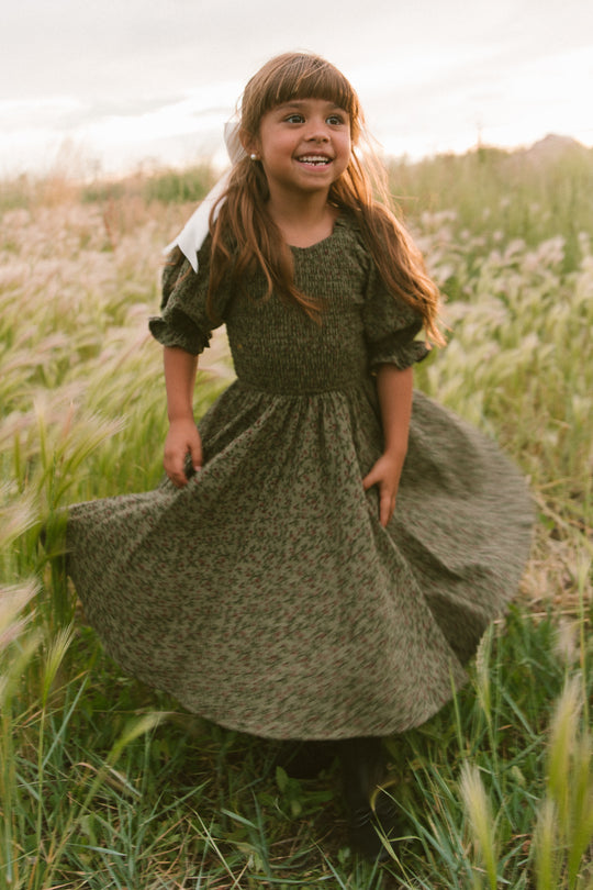 A young girl with long brown hair in a ribbon smiles and spins in the Ivy City Co Mini Teagan Dress in Woodland Floral among tall grass in a sunlit field.